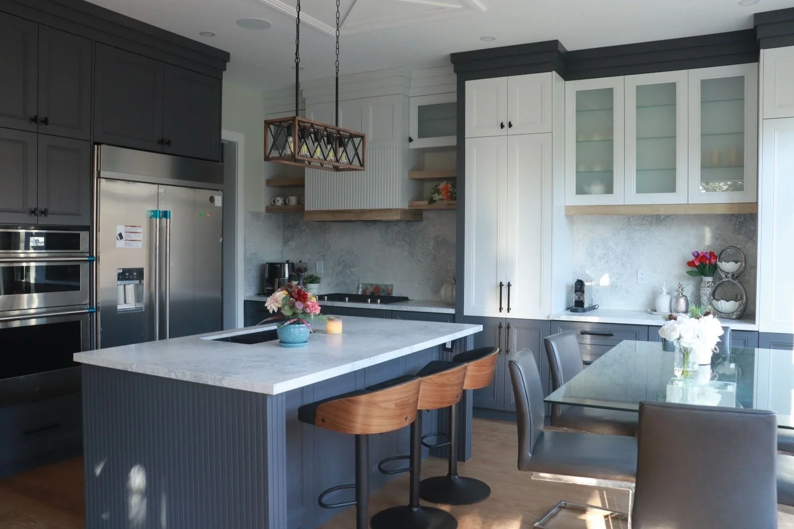 Modern kitchen with gray and white cabinets, stainless steel appliances, a central island with a white marble countertop, wooden seating, and a dining table with chairs. Decor includes flowers and candles.