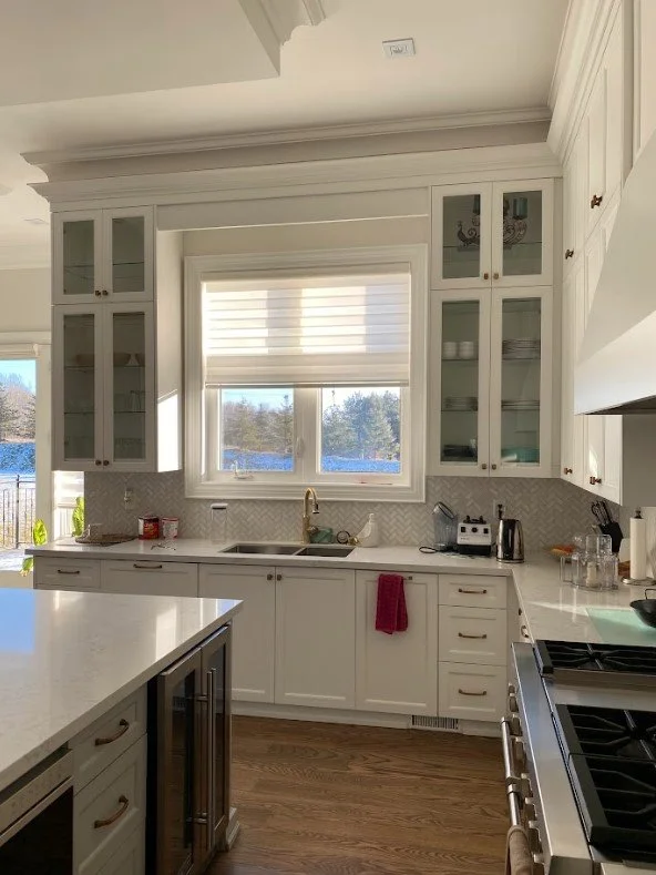 Kitchen with white cabinets, a window with a blind, and a view of a lake and trees outside. Countertops with a red towel, small appliances, and various kitchen items.