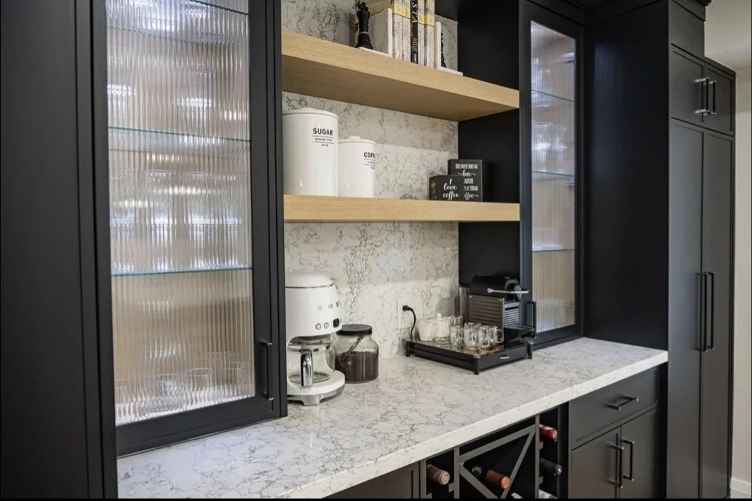 Kitchen counter with black cabinetry, a coffee maker, a coffee machine, glassware, and open shelves with jars labeled sugar and coffee.