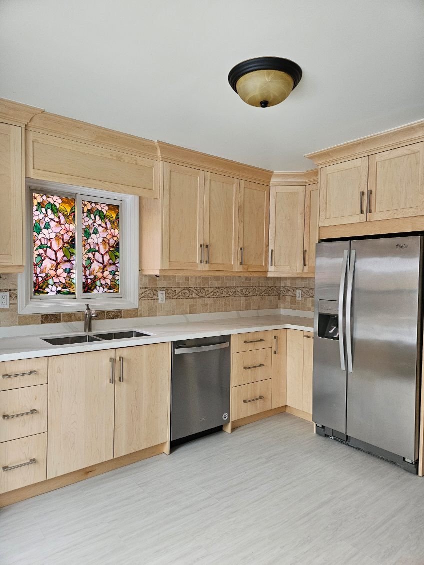 A modern kitchen with light wood cabinets, a stainless steel refrigerator, dishwasher, and a double sink under a stained glass window featuring pink flowers and green leaves.