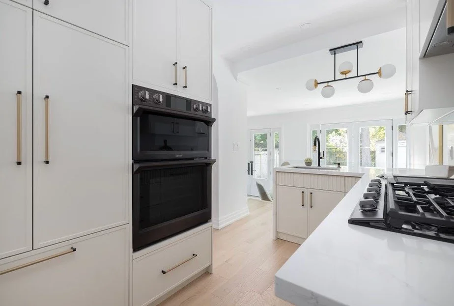 Modern white kitchen with black double oven, white cabinets, and a gas stove on a white countertop. Large windows and a ceiling light fixture are visible in the background.