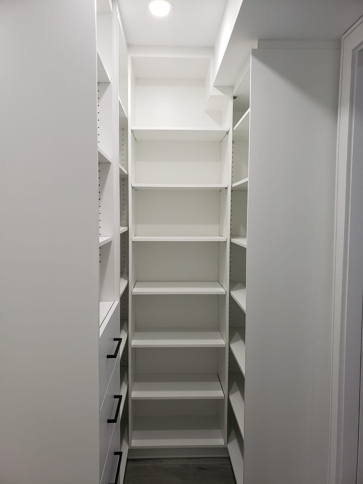 Empty white walk-in closet with multiple shelves and drawers, overhead lighting, and a dark wood floor.