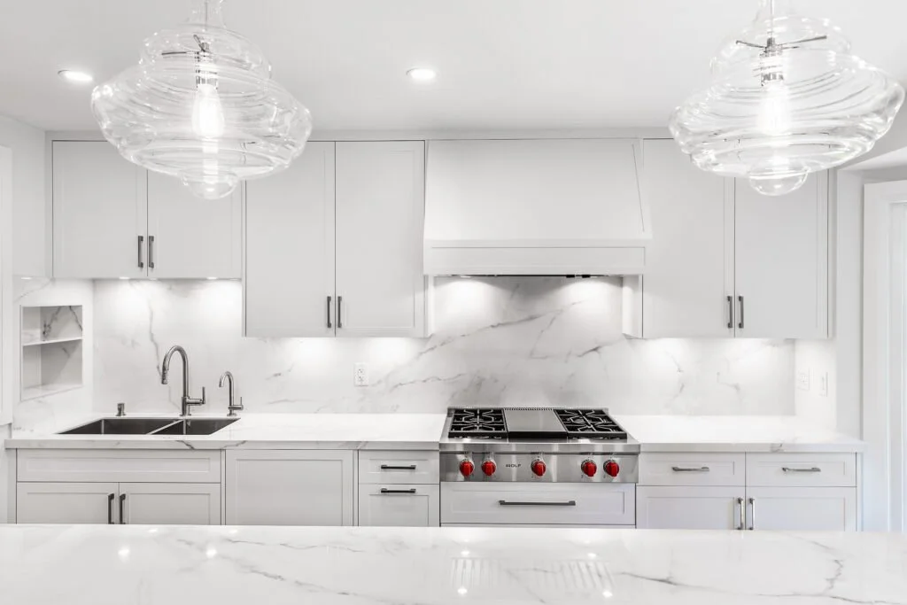 Modern kitchen with white cabinets, marble backsplash, and countertop. Stainless steel stove with red knobs. Overhead glass pendant lights. Double sink with chrome faucets.