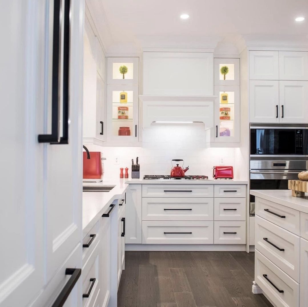 White kitchen with black handles, red accents including a kettle and toaster, with cabinets featuring built-in shelves and a microwave oven, dark hardwood floor.