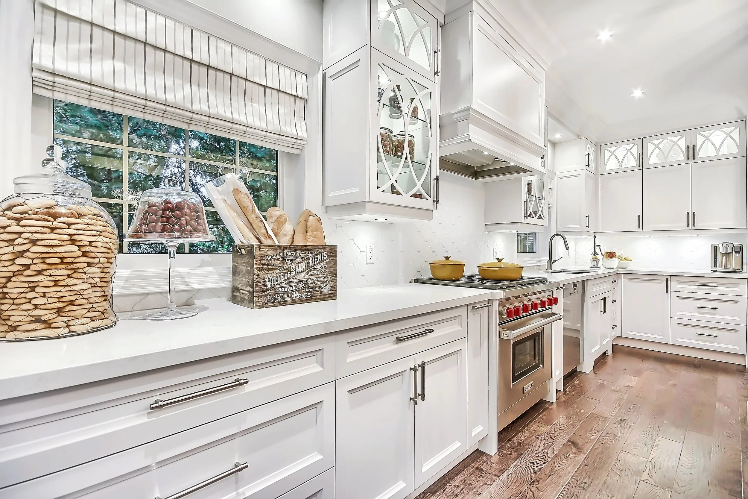 A bright white kitchen with white cabinets, a white countertop, and a window with blinds. The countertop has glass jars filled with cookies and candies, a wooden crate labeled 'VILLE DES SAINT DENIS,' yellow pots on the stove, and small appliances. T