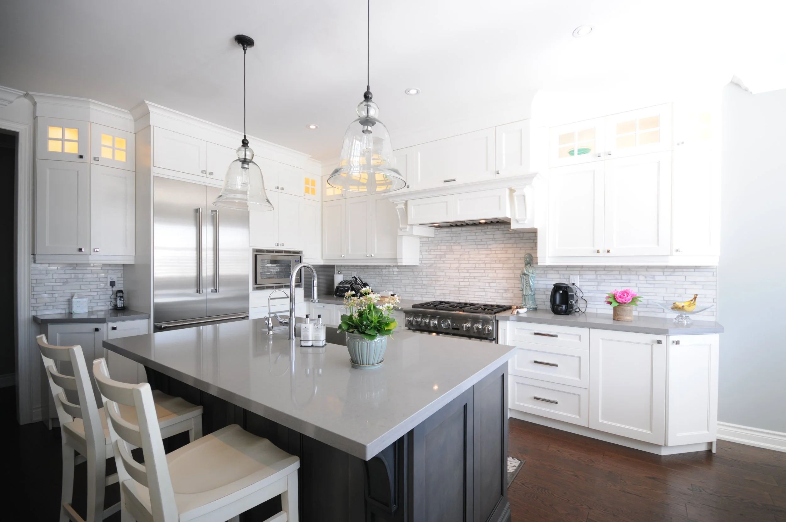 A modern kitchen with white cabinets, a kitchen island with a light-colored countertop, and stainless steel appliances. There are two glass pendant lights hanging above the island, a stove with an exhaust hood, a microwave, a refrigerator, and small 