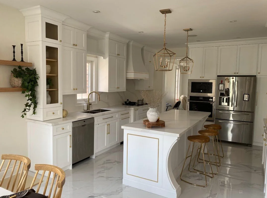 Modern white kitchen with marble floors, gold accents, and a curved island with four wooden bar stools. There are two geometric pendant lights hanging overhead.