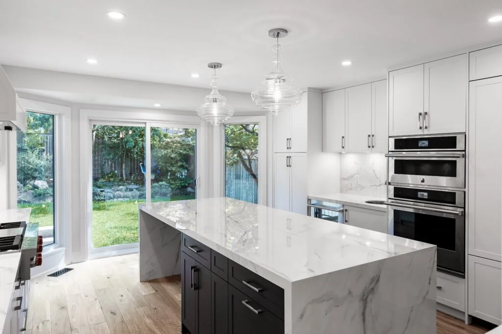 Modern white kitchen with marble countertops, large island, stainless steel appliances, and sliding glass doors to a backyard.