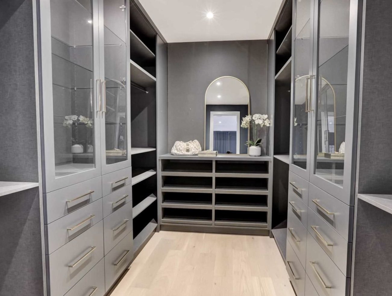 Modern walk-in closet with dark gray walls, light wood flooring, and built-in storage with glass-front cabinets, open shelves, and drawers. A mirror with a gold frame is on the back wall, flanked by two vases of white flowers, and a small stack of books sits on the dresser.