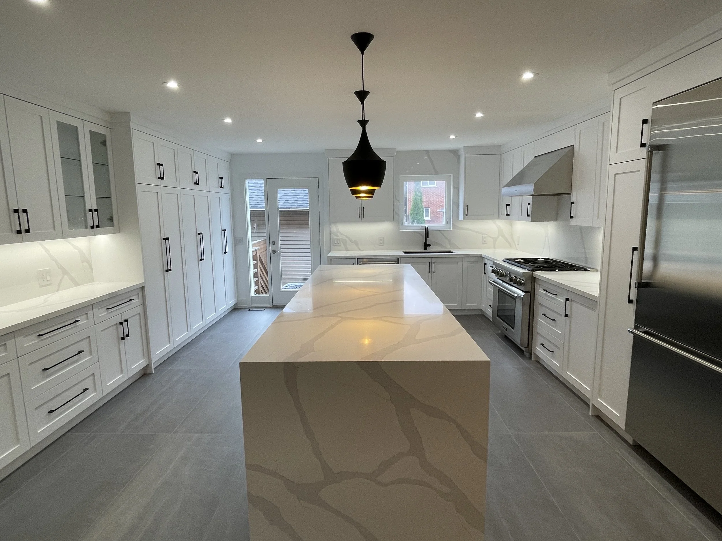 Modern white kitchen with a large central island, black hardware cabinets, stainless steel appliances, and a black pendant light fixture