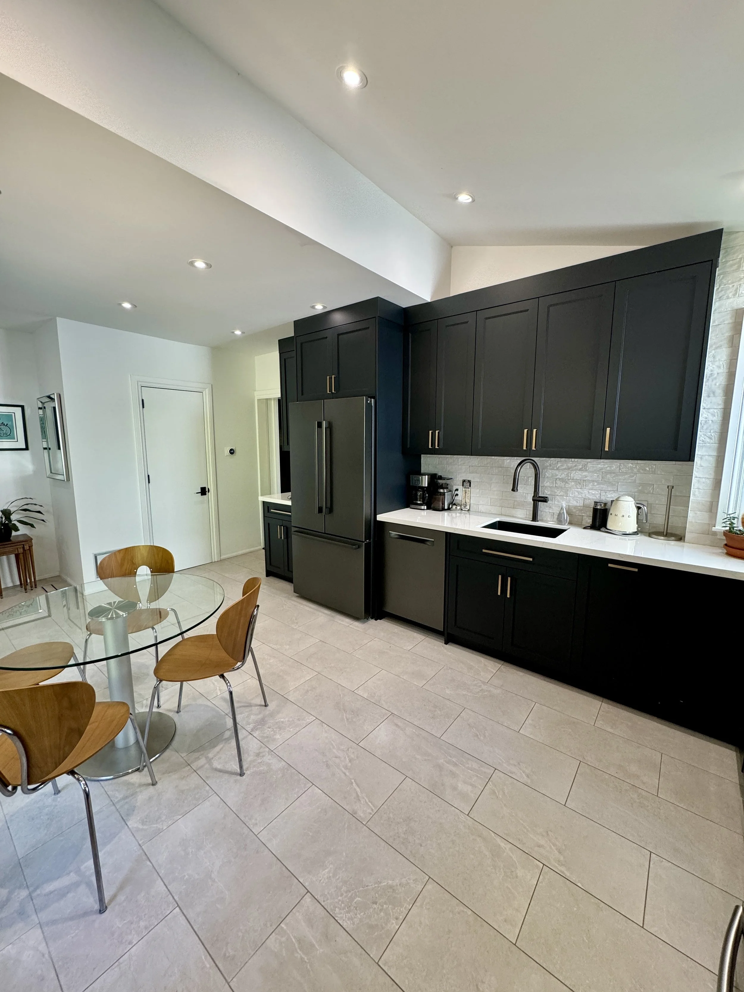 Modern kitchen with black cabinets, white countertop, stainless steel refrigerator, coffee maker, kettle, and a round glass dining table with wooden chairs. Tiled floor and recessed lighting.
