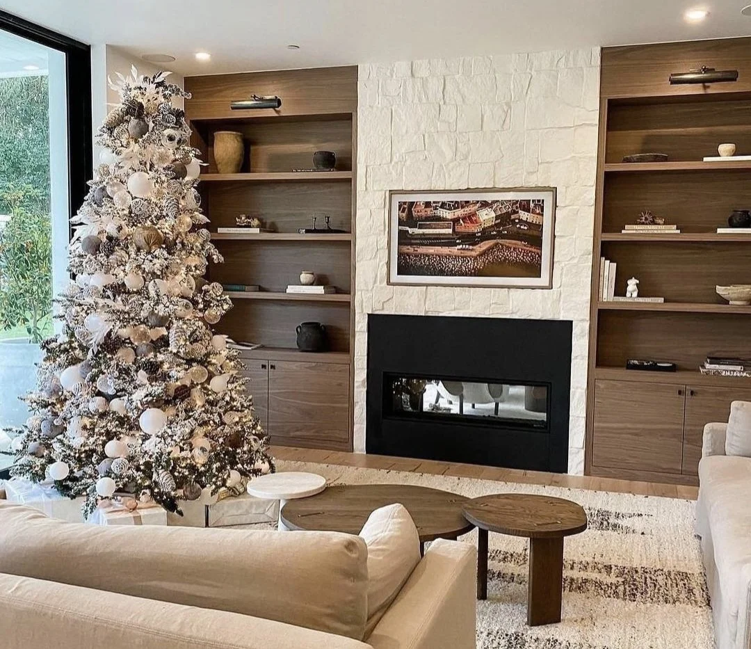 Living room decorated for Christmas with a white Christmas tree, beige sofas, and wooden shelves flanking a fireplace with artwork above.