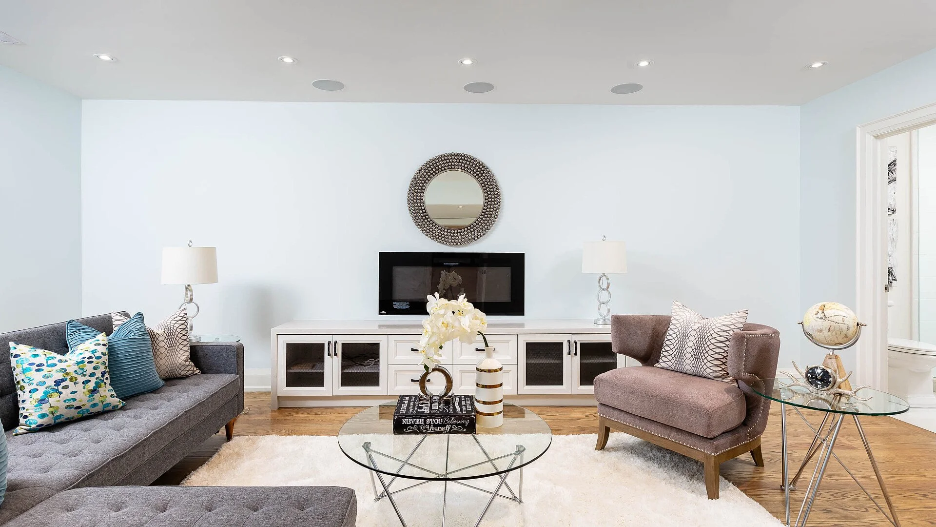 Living room with gray sofa, decorated pillows, glass coffee table with flower vase and books, white TV console, wall mirror, flat-screen TV, side table with globe, armchair, and a white rug.