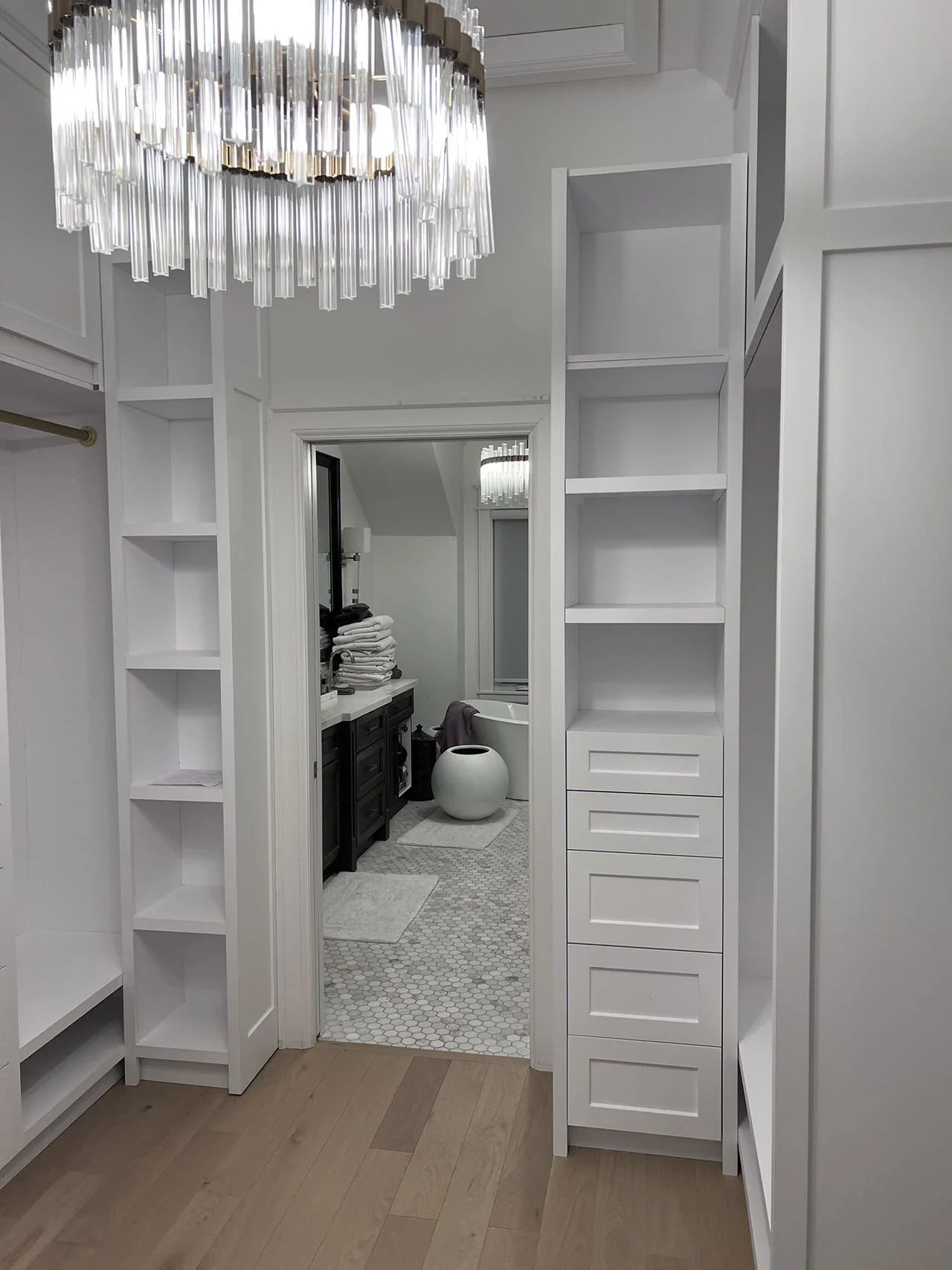 A walk-in closet with white shelving, a chandelier overhead, and a door leading to a bathroom with a vanity, towels, and a bathtub.