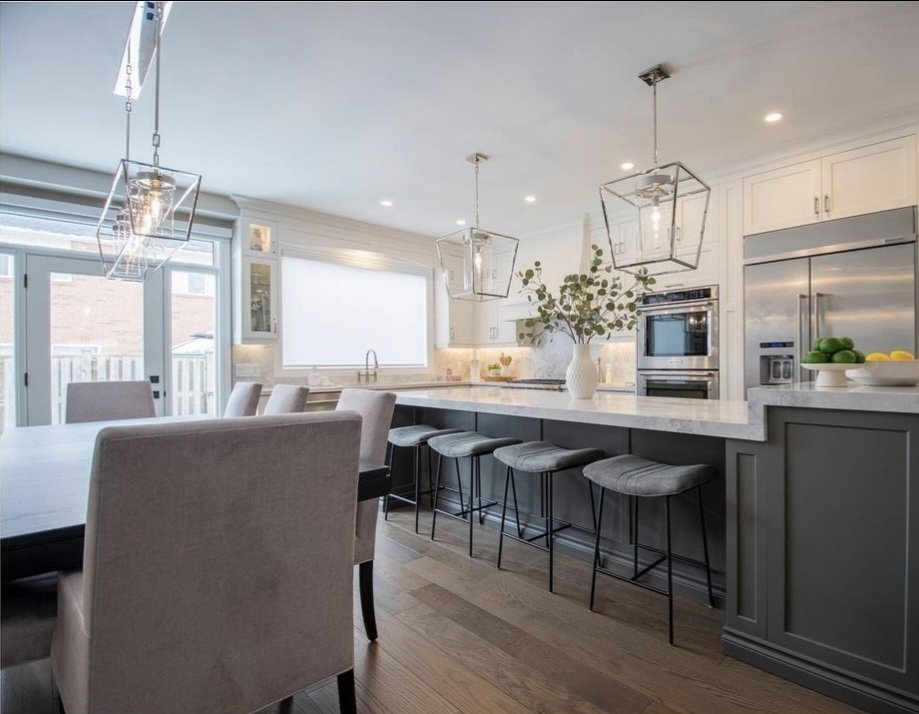 Modern kitchen with white cabinets, gray island with bar stools, stainless steel refrigerator, decorated with a vase of greenery, and illuminated by geometric pendant lights.