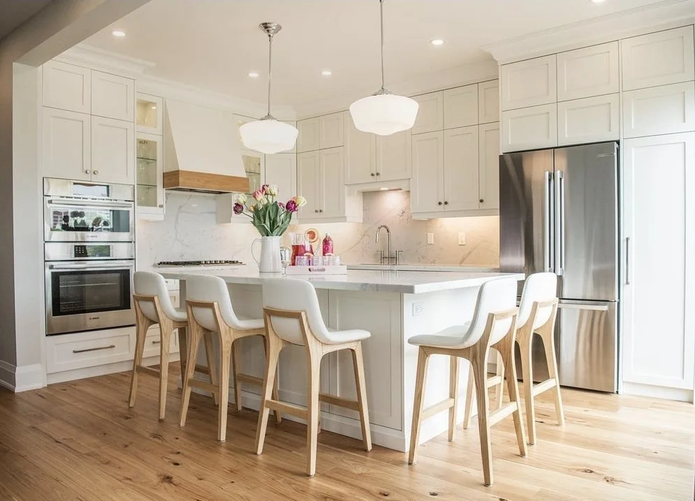 Modern white kitchen with marble countertop island, six white and wood barstools, stainless steel refrigerator, built-in oven, and floral centerpiece.