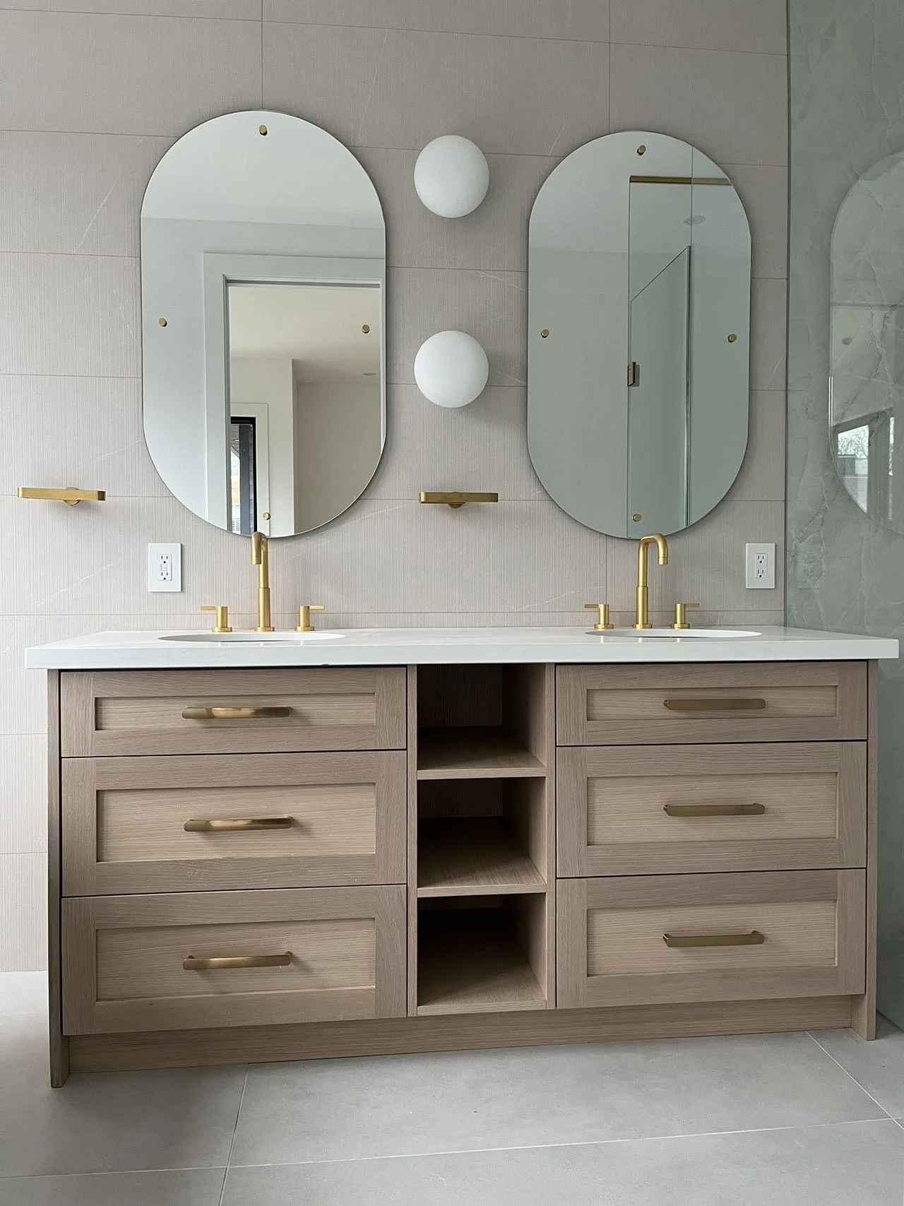 A modern bathroom with a double vanity featuring a white countertop, two gold faucets, six drawers and three open shelves, two oval mirrors with gold accents, and two round white wall sconces.