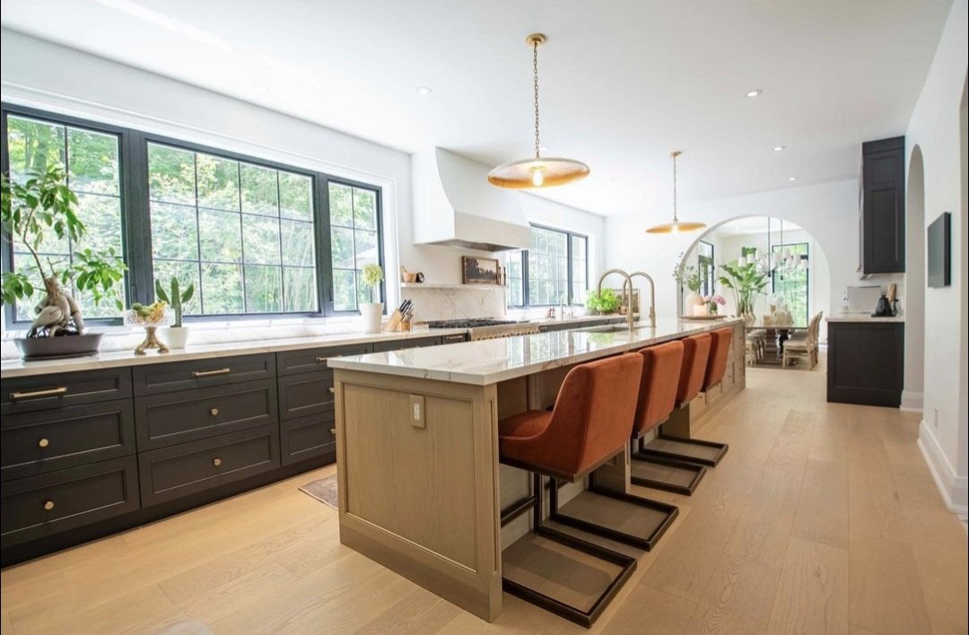 Modern kitchen with large windows, a central island with brown barstools, dark cabinetry, and light hardwood floors.