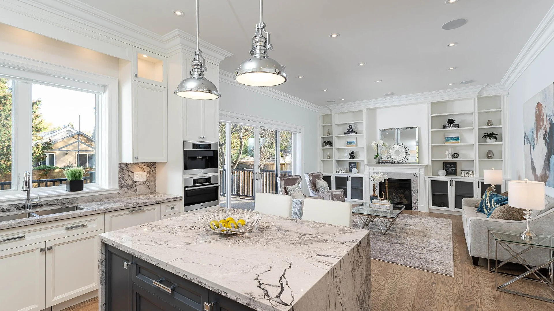 Open-concept kitchen and living room with white cabinetry, marble countertops, stainless steel appliances, built-in shelves, fireplace, large windows, and modern decor.
