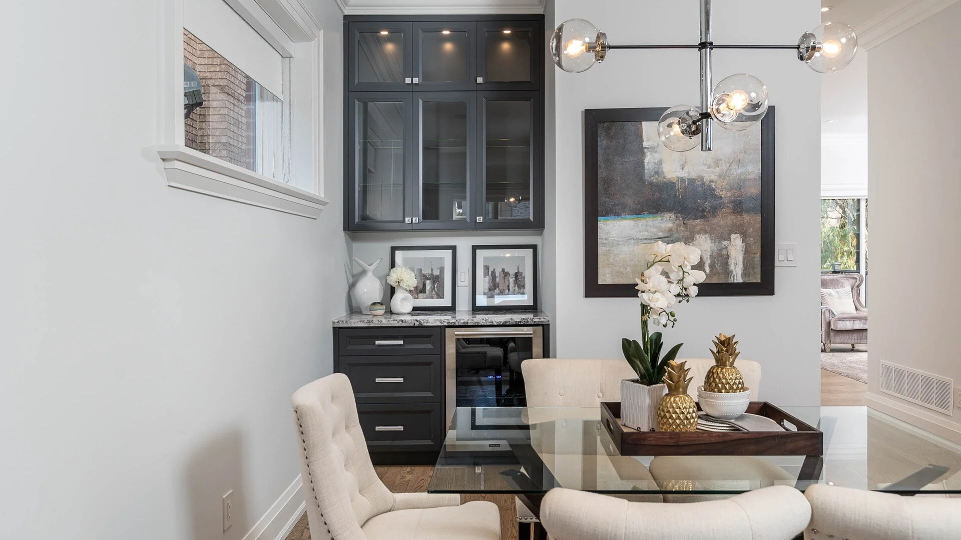 A modern dining room with beige upholstered chairs around a glass table, decorated with white orchids and gold pineapple ornaments, a large abstract painting on the wall, and a black cabinet with glass doors and framed pictures below it. There is a l