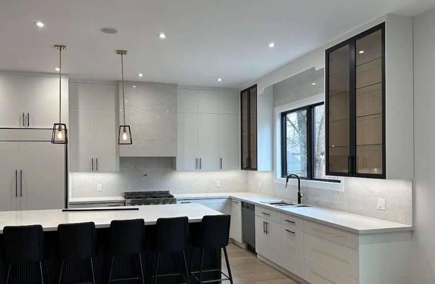 Modern kitchen with white cabinets, marble countertops, black bar stools, pendant lights, and a window above the sink.