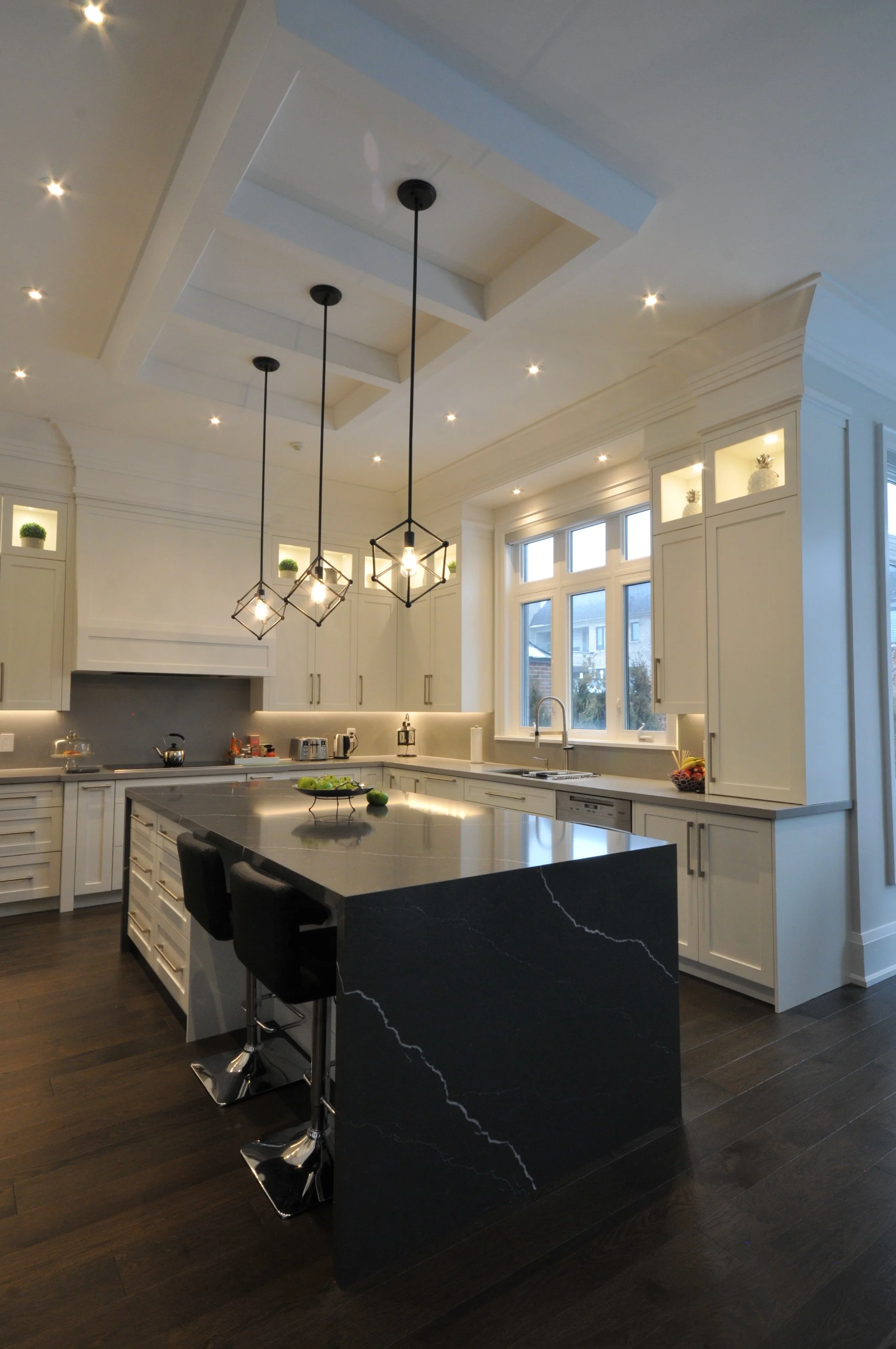 Modern kitchen with white cabinets, black marble island with white veining, pendant lights, large window, dark wood flooring, and small potted plants in built-in shelves.