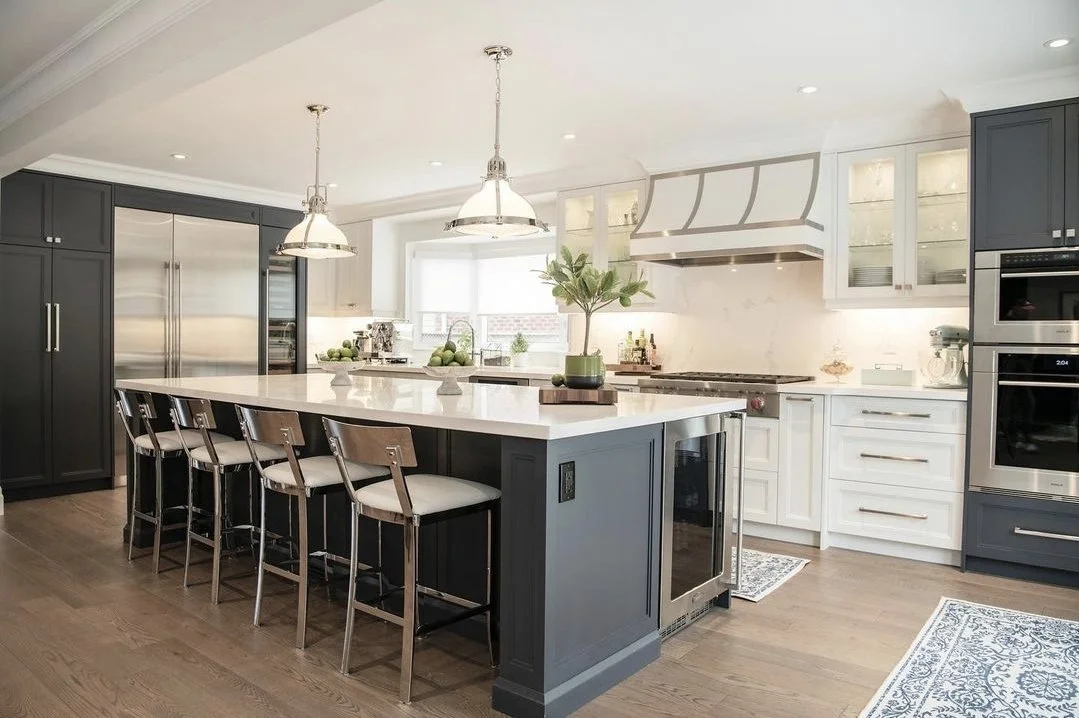 Modern kitchen with white and navy cabinetry, stainless steel appliances, island with seating, pendant and recessed lighting, wooden floor, and decorative plants and fruit accents.