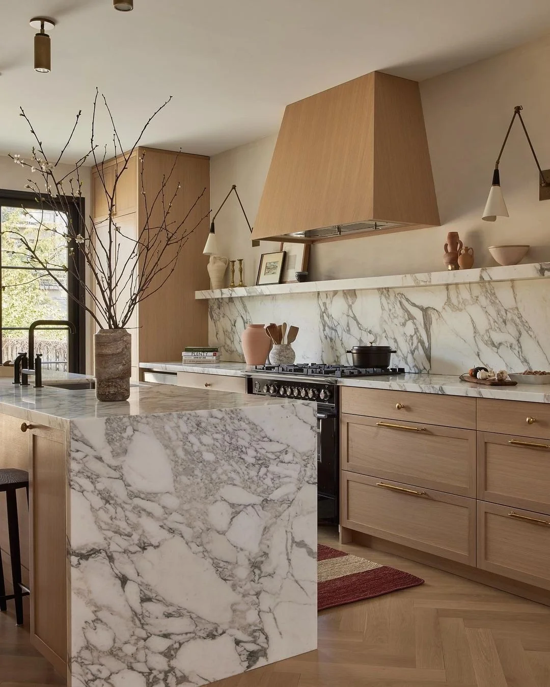 Modern kitchen with marble countertops and backsplash, light wood cabinets, a large vase with branches on the island, and decorative items on the shelf.