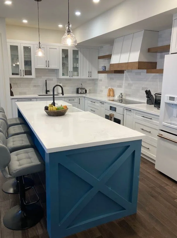 Modern kitchen with white cabinets, a large island with a blue base and white countertop, pendant lights, and wooden floating shelves.