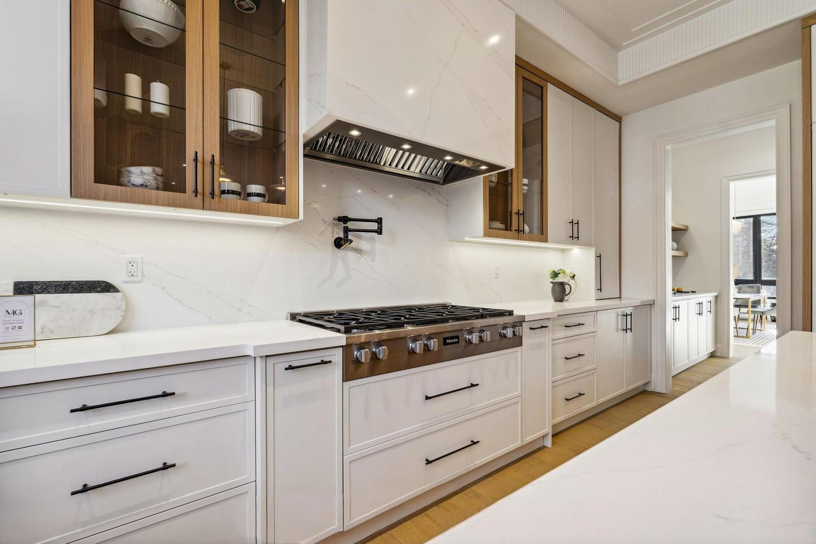 Modern kitchen with white cabinets, marble countertops, and a stainless steel gas range stove. Wooden upper cabinets with glass doors, black hardware, and a marble backsplash. Visible doorway leading to a bright dining area.