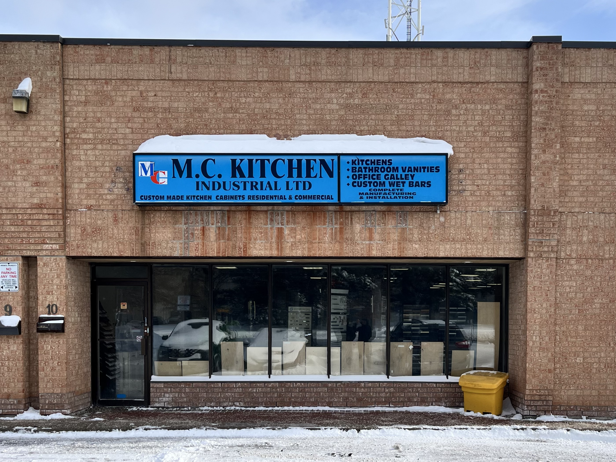Brick building with a blue sign reading 'M.C. KITCHEN INDUSTRIAL LTD' and listing services such as kitchens, bathroom vanities, office gallery, and custom wet bars. The building has large glass windows, a door, and snow on the ground.
