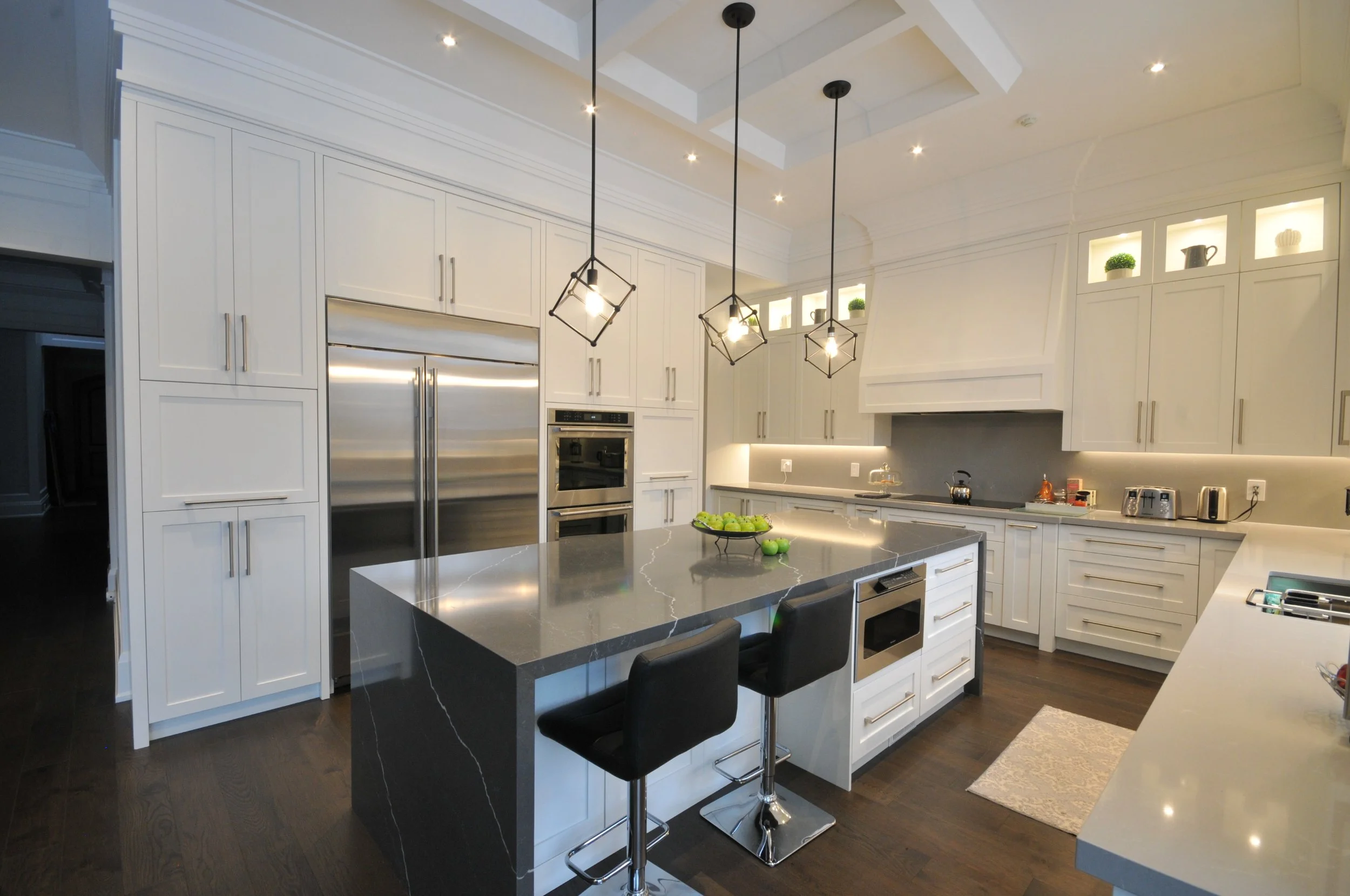 Modern white kitchen with black and gray accents, featuring a large island with a dark gray marble countertop, black bar stools, pendant lighting, built-in appliances, and decorative items displayed in illuminated upper cabinets.