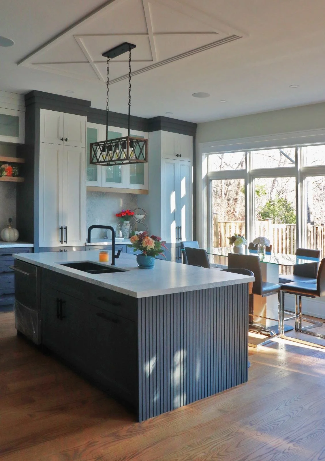 Modern kitchen with white cabinets, a kitchen island with a white countertop and dark base, a black faucet, dining table near large windows, and floral decor.