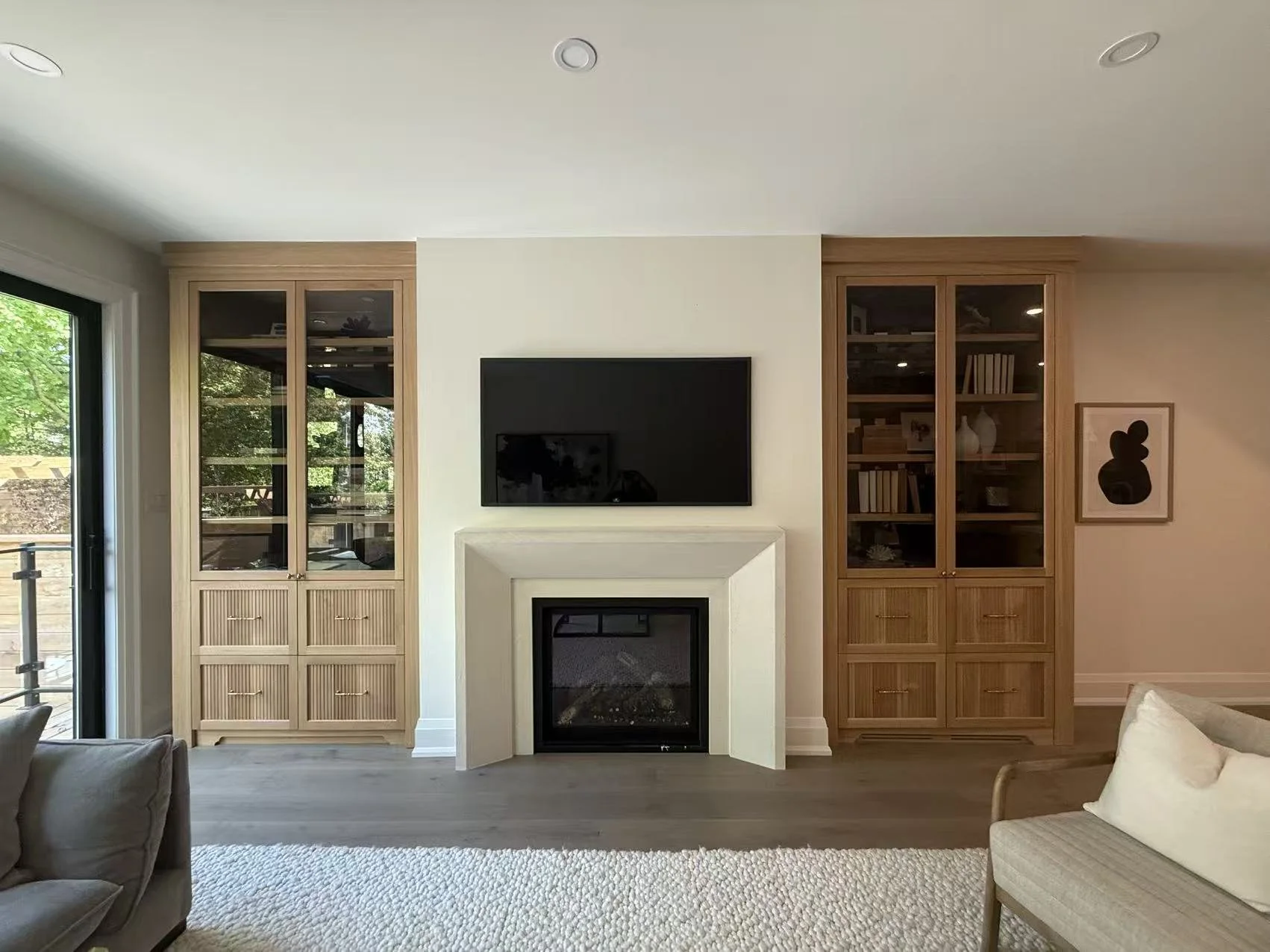 Living room with built-in wooden bookcases flanking a fireplace and a television mounted above the fireplace, a sliding glass door on the left, and a framed abstract art on the right.