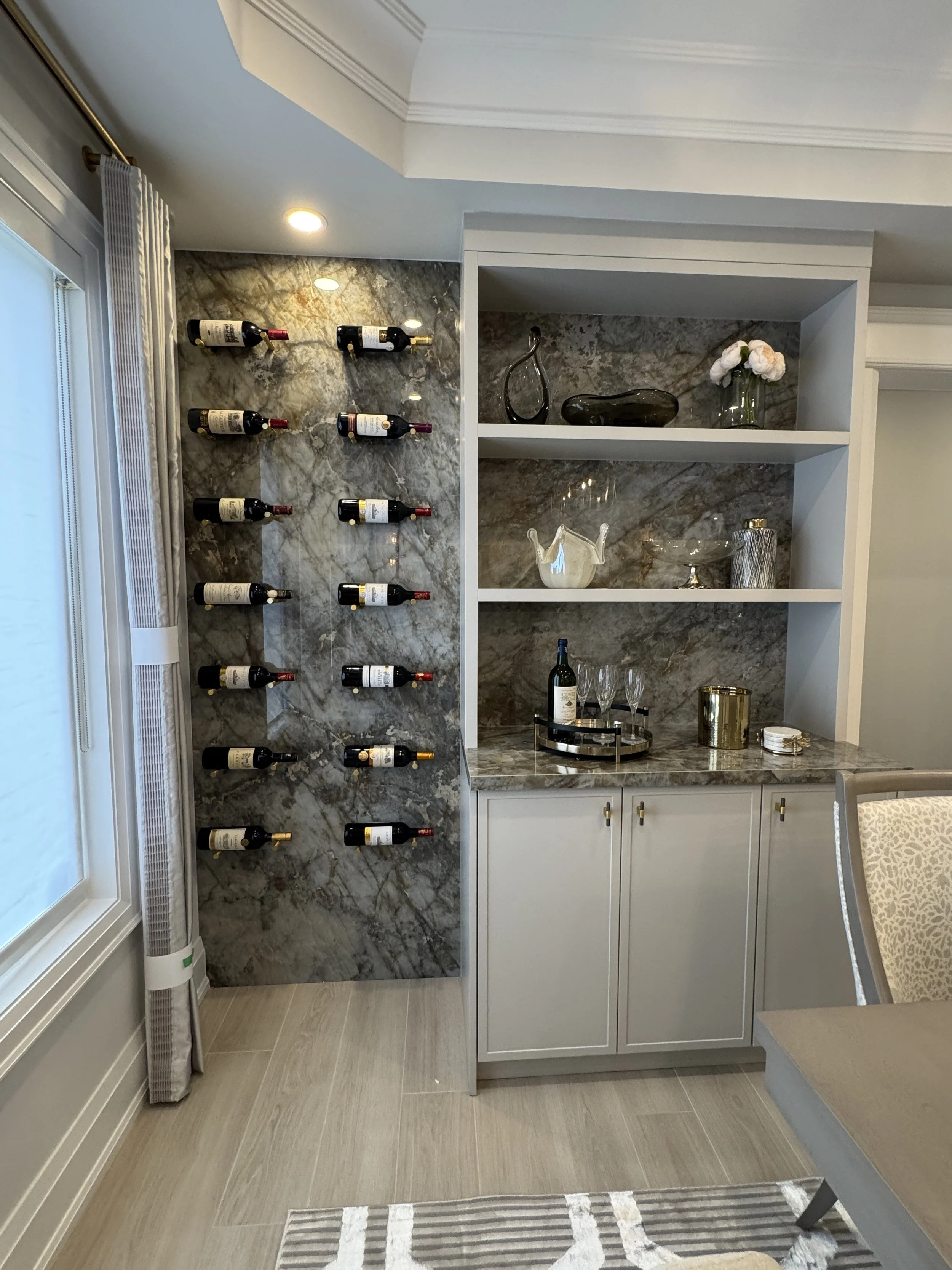 A wine storage wall with multiple wine bottles mounted on a marble background adjacent to a built-in cabinet with decorative items and glassware.