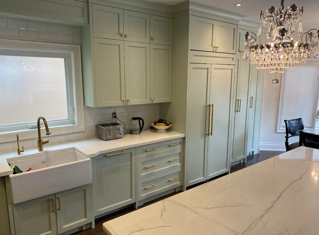 Kitchen with light green cabinets, white countertop, farmhouse sink, a window above the sink, a chandelier hanging from the ceiling, and a bowl of fruit on the counter.