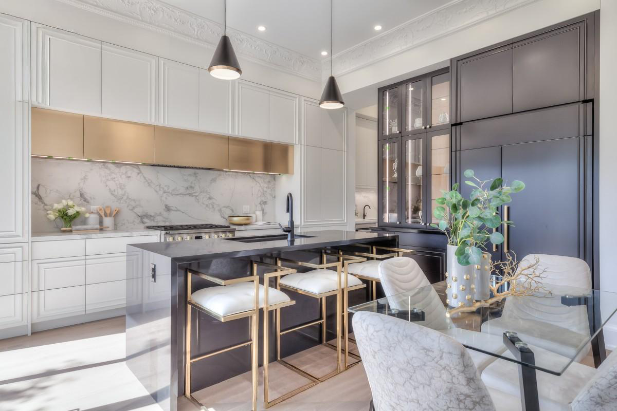 Modern kitchen with black and white cabinetry, marble backsplash, glass-front cabinets, black countertops, and a glass dining table with white and marble chairs, decorated with a vase of greenery.