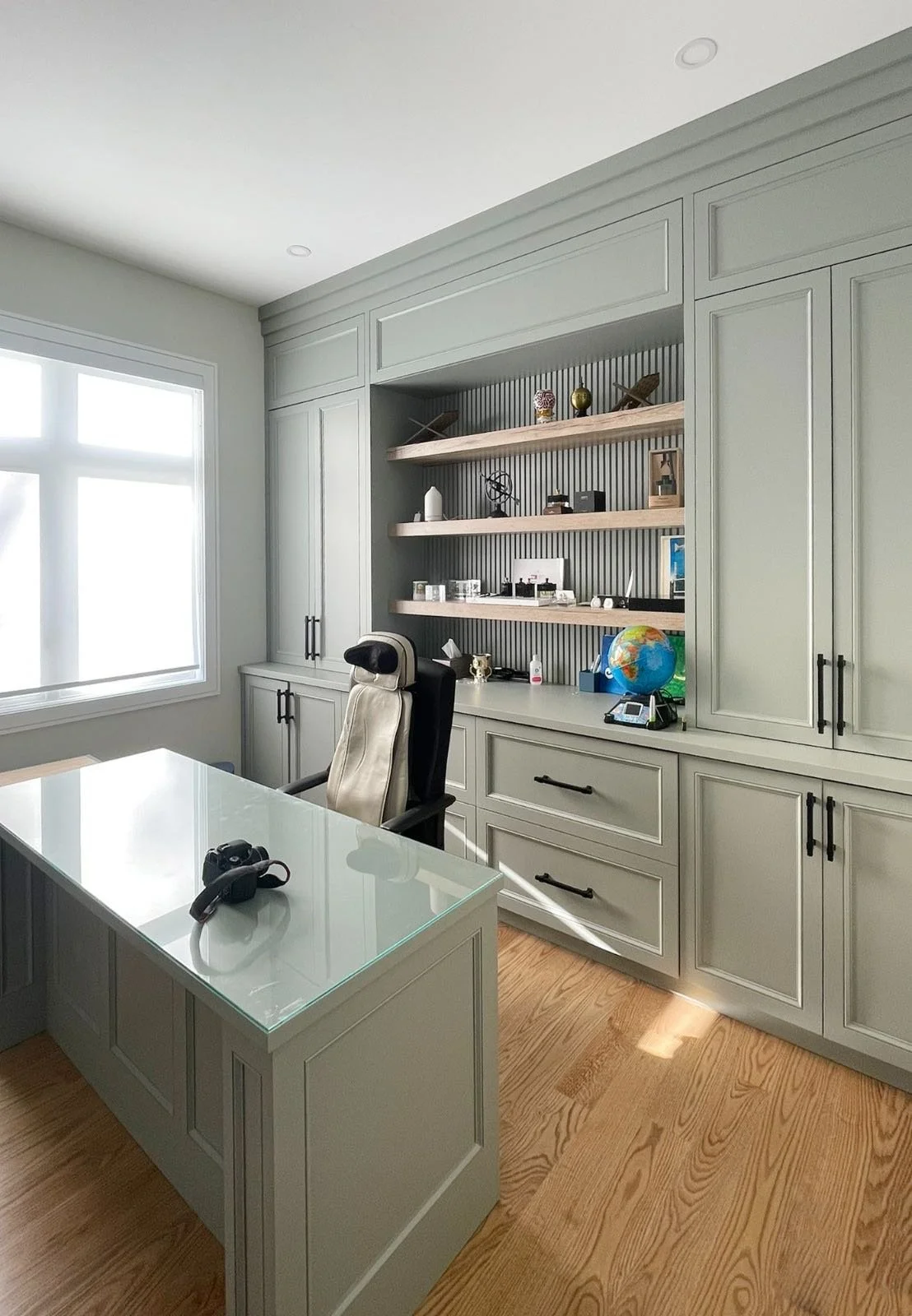 A home office with a large window, gray cabinets, open shelves with decorative items, a desk with a camera, a black and beige office chair, and a globe.