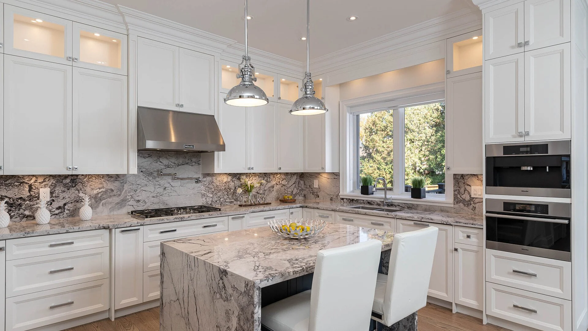 Modern white kitchen with marble countertops and backsplash, stainless steel appliances, and large window with view of trees.