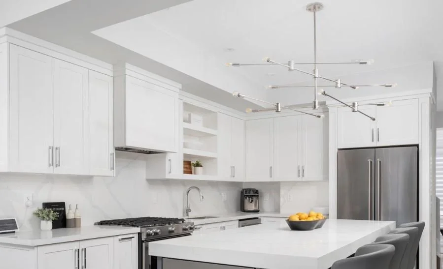 Modern white kitchen with stainless steel appliances and open shelves, featuring a large island with a bowl of lemons and gray chairs.