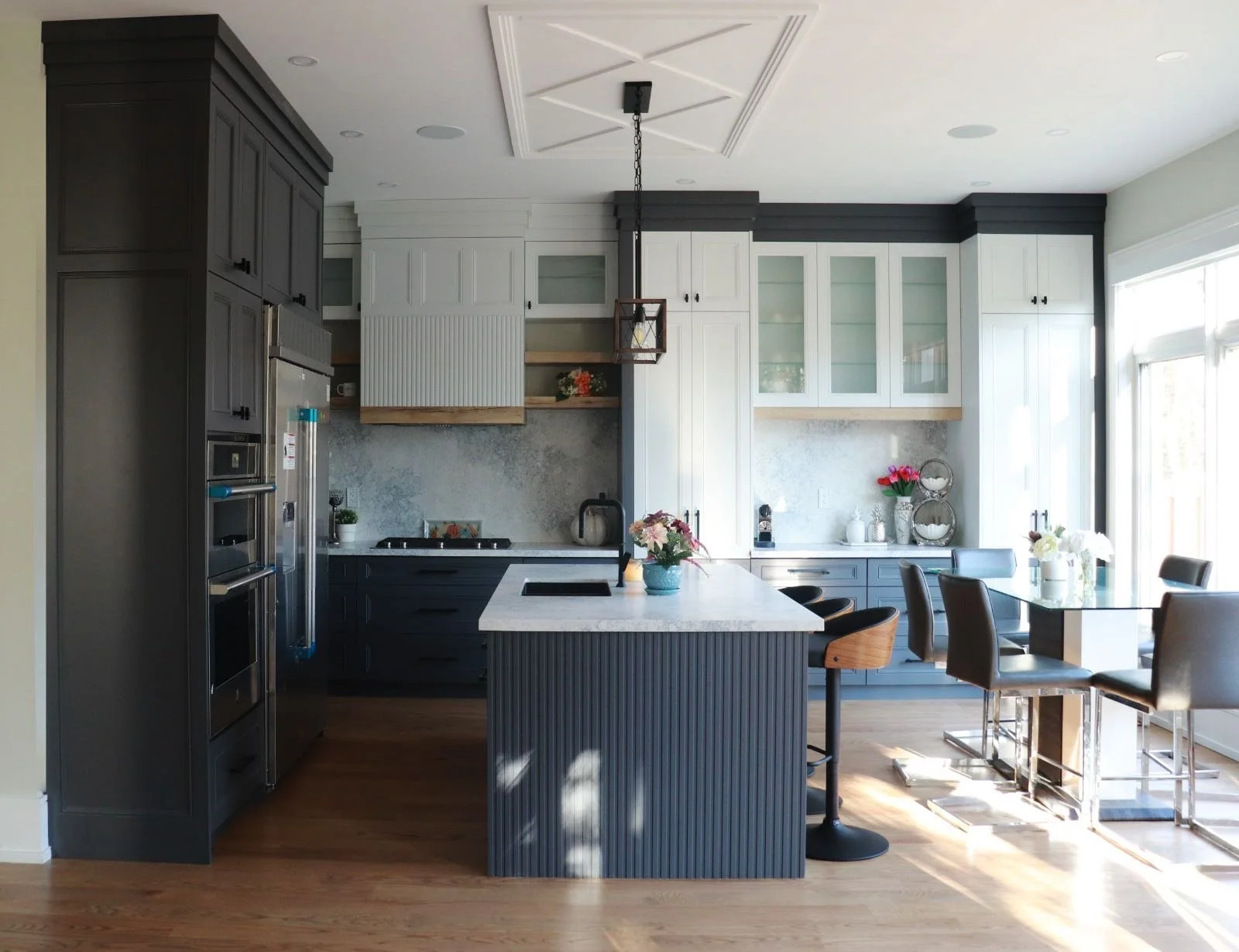 Modern kitchen with gray and white cabinets, a marble island with a potted plant, a dining area with a glass table and black chairs, and large windows letting in natural light.