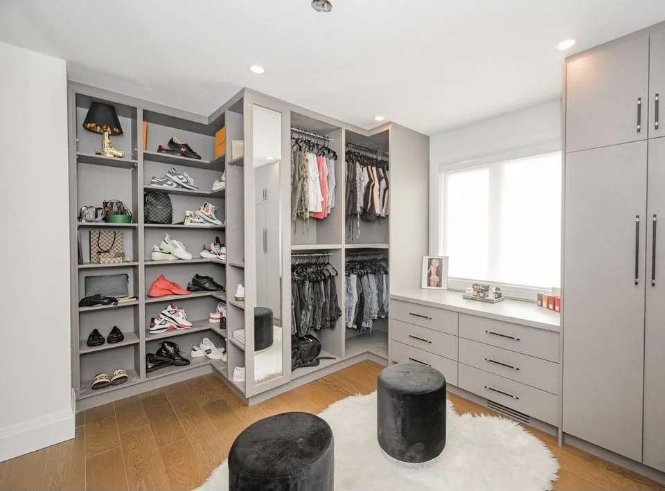 A walk-in closet with gray built-in shelves filled with shoes, handbags, and accessories, alongside hanging clothing. There's a window with white blinds, a dresser below the window with small items on top, and a white fuzzy rug with two black stools 