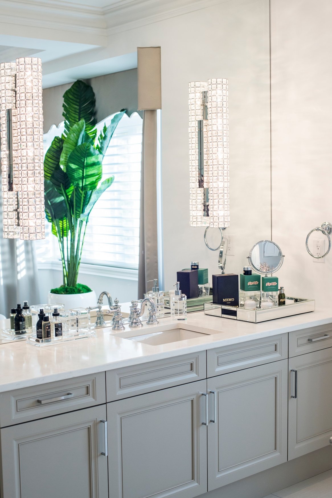 A bathroom vanity with a mirror, decorative lighting, and various skincare and beauty products on the countertop. There is a large potted plant near the window.