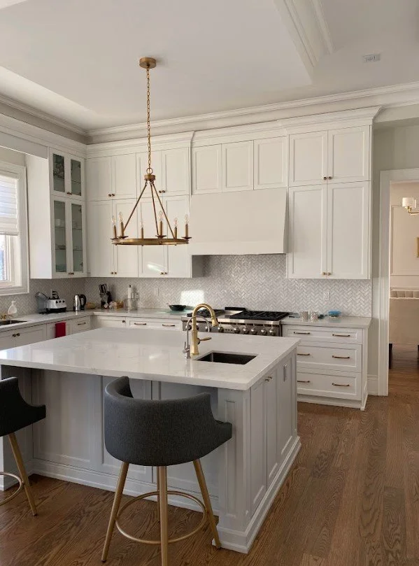 Modern white kitchen with a central island, gold faucet, chandelier, and hardwood floors.