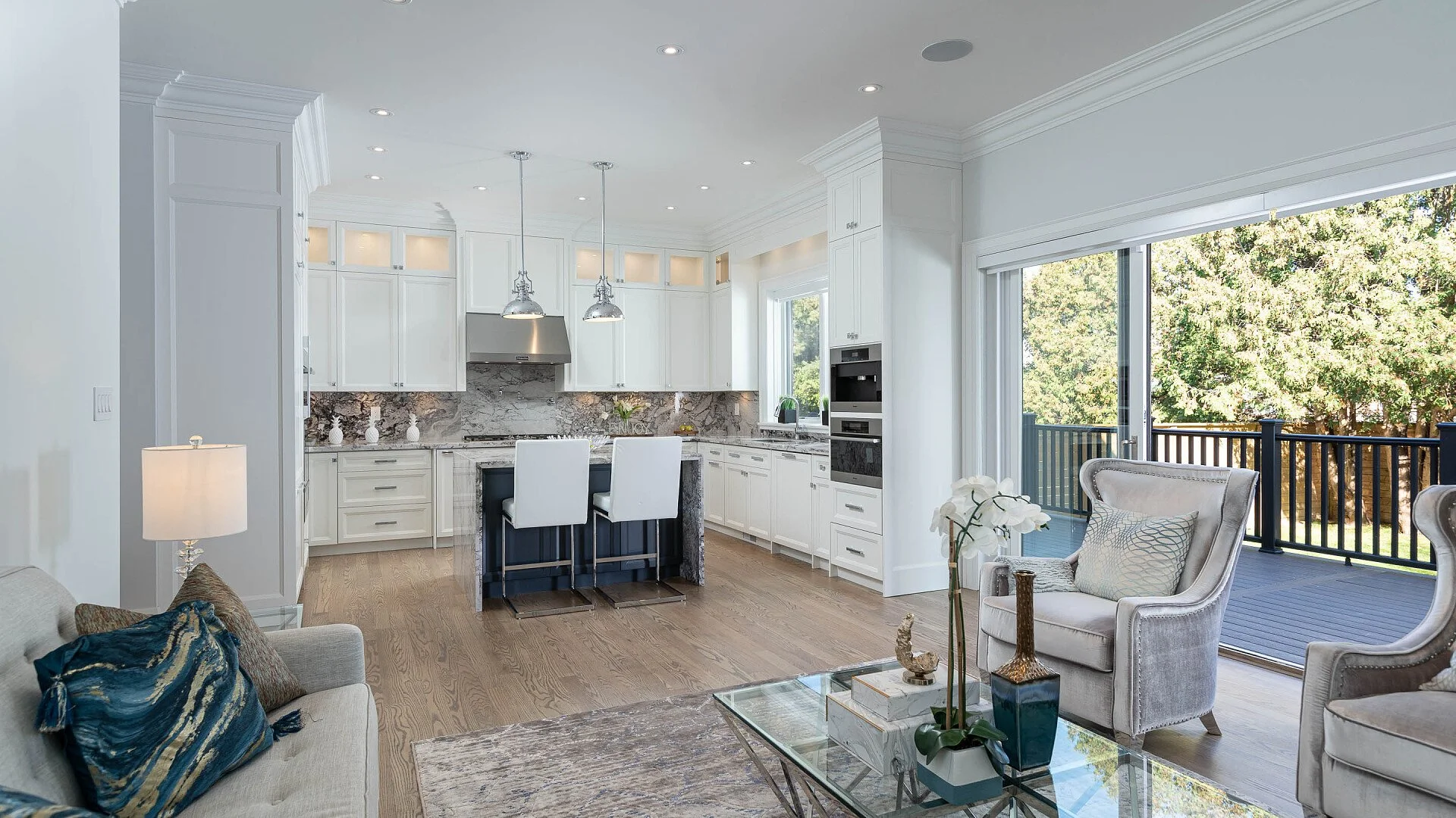 Bright, modern kitchen and living room with white cabinets, stainless steel appliances, marble backsplash, and large glass sliding doors opening to a wooden deck surrounded by trees.