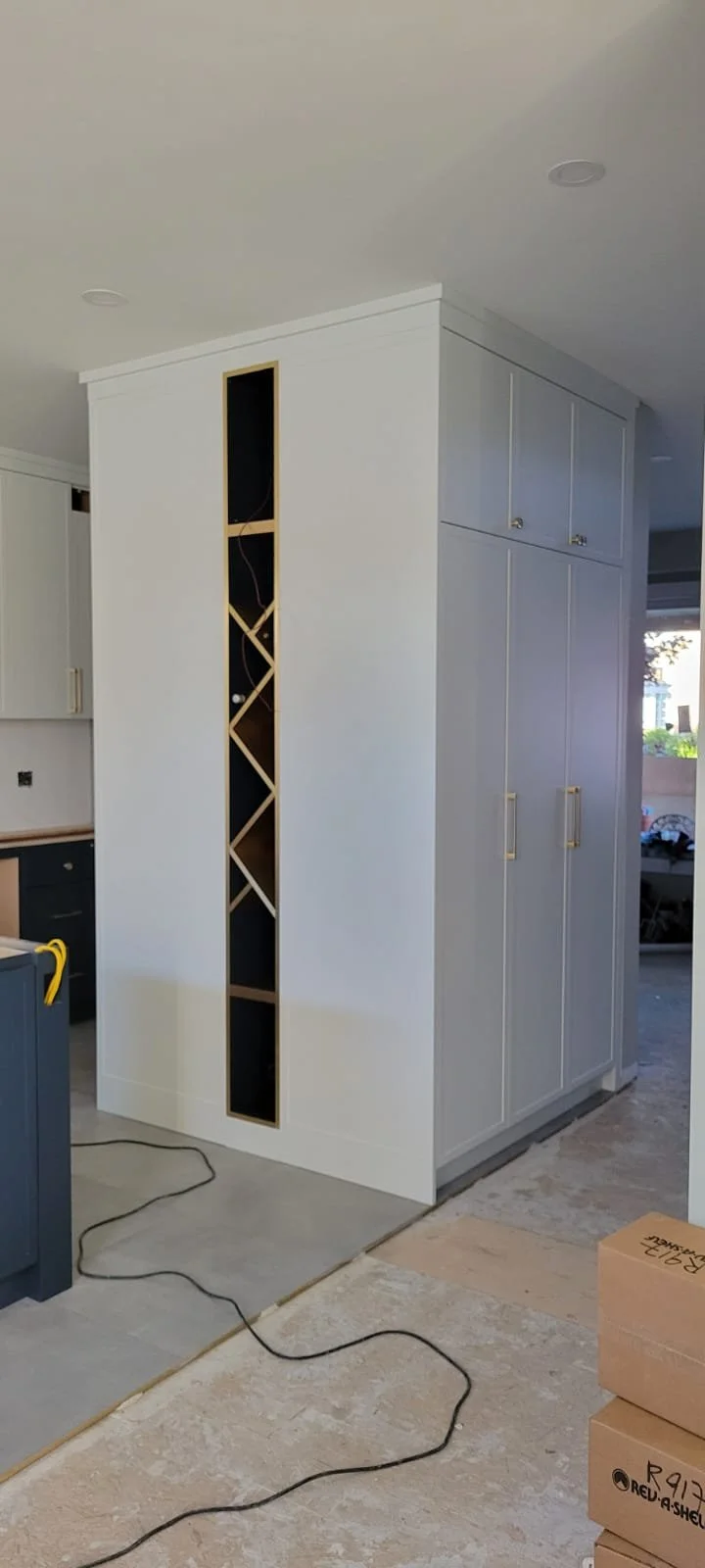 Partially finished kitchen with white cabinets, a corner wine rack, and a black and gold-themed cabinet. Construction tools and boxes are on the unfinished floor.