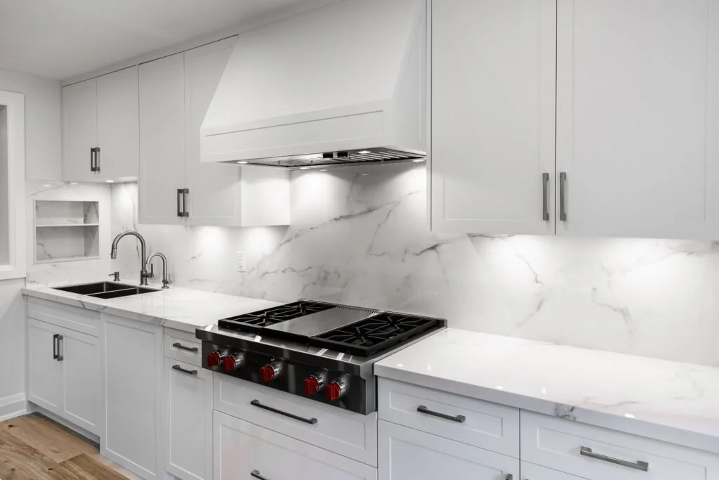Modern white kitchen with marble backsplash and countertops, black stove with red knobs, under-cabinet lighting, and a double sink.