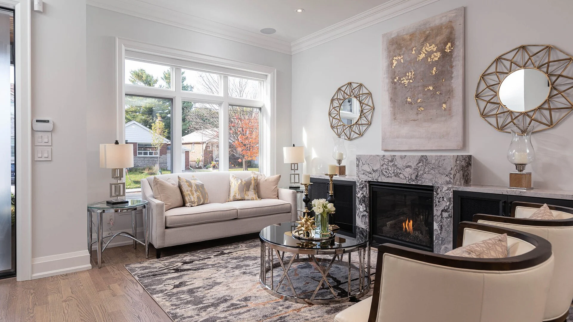 Living room with a white sofa, beige pillows, a glass coffee table with decorative items, a fireplace with a marble surround, and large windows showing an outdoor scene with trees and houses. There are two armchairs with beige cushions, wall mirrors, and abstract wall art.