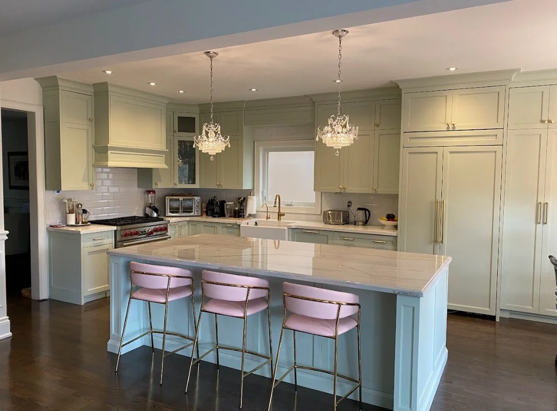 Modern kitchen with pastel yellow cabinets, white subway tile backsplash, marble island, pink chairs, and two chandeliers.