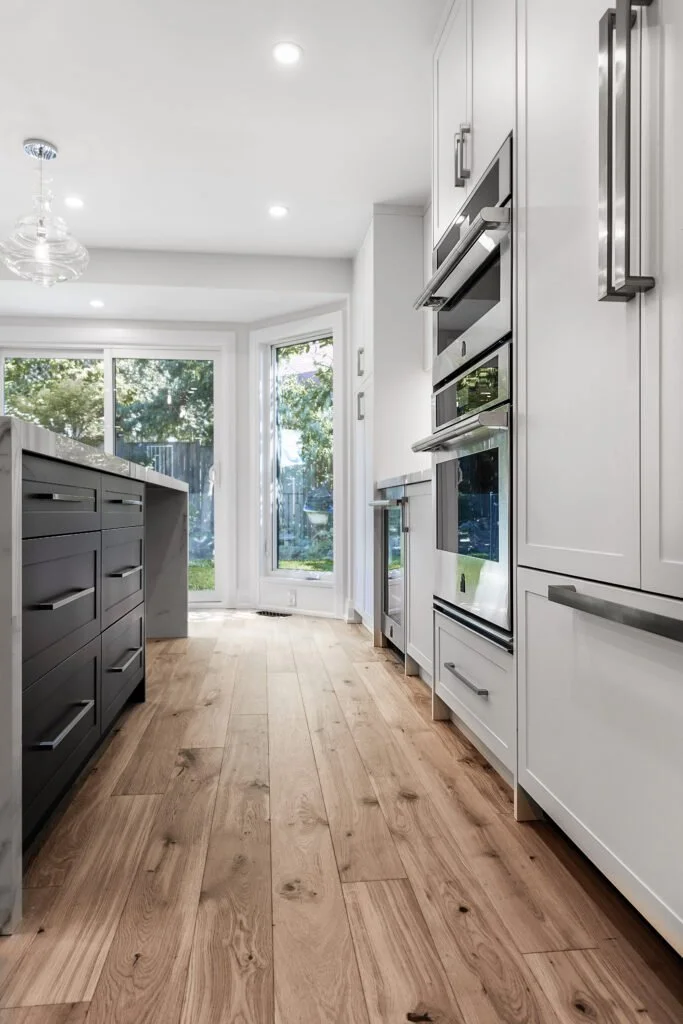 Modern kitchen with light-colored cabinets, stainless steel appliances, wood flooring, and a large window with a view of the backyard.